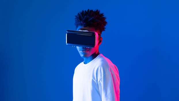 Surprised young African American looking through VR holding interesting object hologram on pink blue neon wall at metaverse world connecting digital futuristic technology virtual reality. Contrivance.