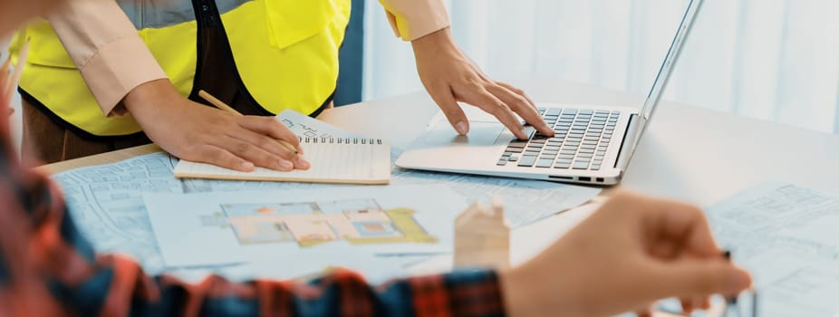 Cropped image of professional engineer team working on blueprint while his coworker working on laptop at meeting table with blueprint and wooden block scattered around. Closeup. Delineation.