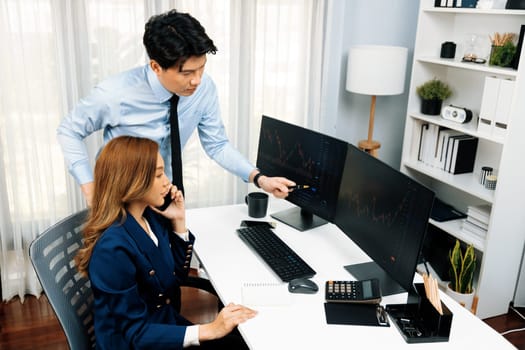 Smiling Asian businesswoman calling to broker dynamic stock exchange database with businessman partner pointing to screens comparing profit value online application program at home office. Infobahn.