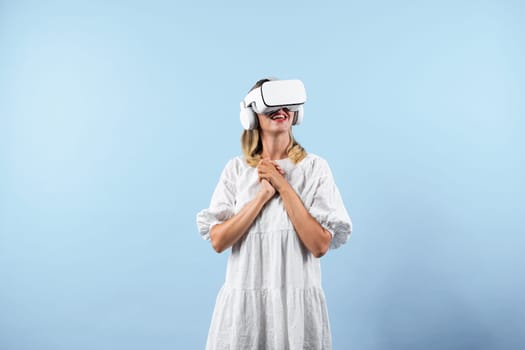Happy girl enjoy looking at nature view hologram in VR glasses while connect at metaverse or visual reality world. Caucasian woman with white pajamas while standing at blue background. Contraption.