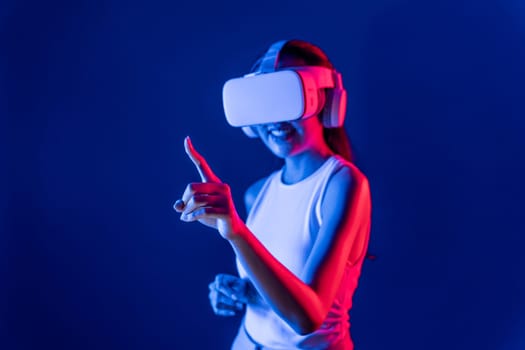 Smart female stand with surrounded by cyberpunk neon light wear VR headset connecting metaverse, futuristic cyberspace community technology. Woman using finger pointing virtual object. Hallucination.