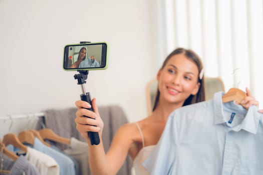 Young woman making fashion video content for social media. Blogger smiles to camera using selfie stick and light ring while making persuasive online clothing sell to audience or followers. Unveiling