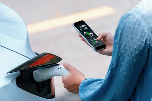 Hand insert EV charger plug into electric vehicle to recharge EV car, battery status display on smartphone EV application. Future alternative clean and sustainable energy for transportation. Perpetual