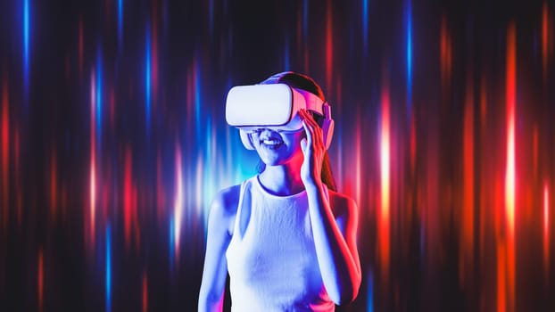 Smart female standing surrounded by neon light wearing VR headset connecting metaverse, future cyberspace community technology. Elegant woman looking faraway and smiling satisfactorily. Hallucination.