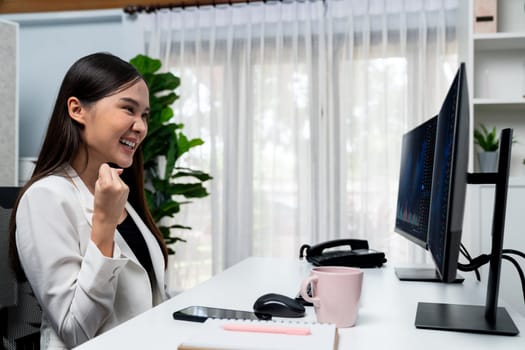Successful young Asian businesswoman earning highest profit with fist up in exchange stock market on dynamic business graph on pc screens at modern office, analyzing financial technology. Stratagem.