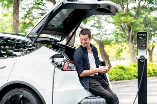 Man using smartphone online banking application to pay for electric car battery charging from EV charging station during vacation holiday road trip at national park or summer forest. Exalt