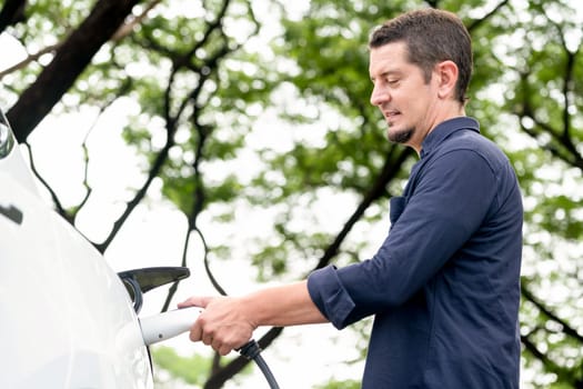 Man recharging battery for electric car during road trip travel EV car in natural forest or national park. Eco friendly travel during vacation and holiday. Exalt