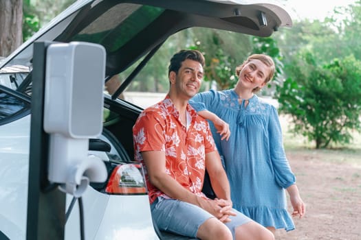 Road trip vacation with electric vehicle, lovely couple sitting on trunk and recharge EV car with green and clean energy. Nature and travel with eco-friendly car for sustainable environment. Perpetual