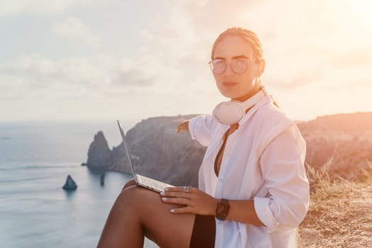 Digital nomad, woman in the hat, a business woman with a laptop sits on the rocks by the sea during sunset, makes a business transaction online from a distance. Freelance, remote work on vacation.