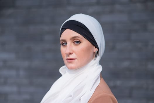 Portrait of a young blue-eyed woman in a hijab against a gray brick wall