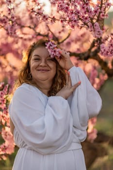 Woman blooming peach orchard. Against the backdrop of a picturesque peach orchard, a woman in a long white dress enjoys a peaceful walk in the park, surrounded by the beauty of nature