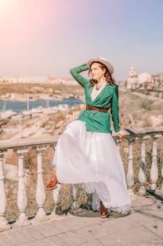 Woman walks around the city, lifestyle. A young beautiful woman in a green jacket, white skirt and hat is sitting on a white fence with balusters overlooking the sea bay and the city