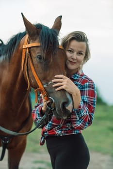 Happy blonde with horse in forest. Woman and a horse walking through the field during the day. Dressed in a plaid shirt and black leggings