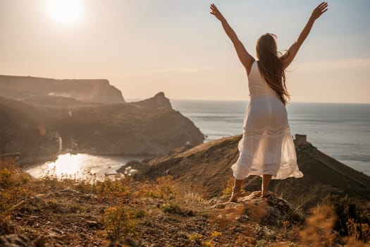 A woman in a white dress stands on a rocky hill overlooking the ocean. She is smiling and she is happy