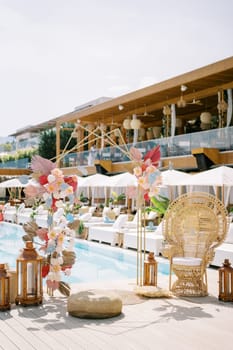 Colorful wedding arch stands near a wicker chair by the pool with double sun loungers under parasols. High quality photo