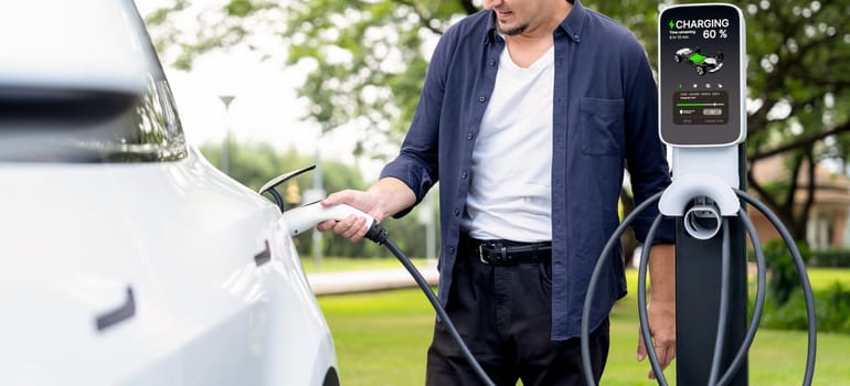 Man recharge EV electric vehicle's battery from EV charging station in outdoor green city park scenic. Eco friendly urban transport and commute with eco friendly EV car travel. Exalt