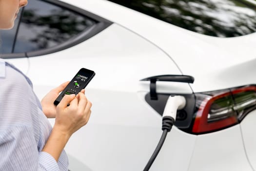 Young woman using smartphone online banking application to pay for electric car battery charging from EV charging station during vacation holiday road trip at national park or summer forest. Exalt