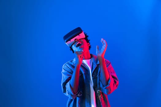 Young African American singer using through VR glasses enjoy singing song with karaoke meta application on blue neon light wall background in virtual reality metaverse fantasy technology. Contrivance.