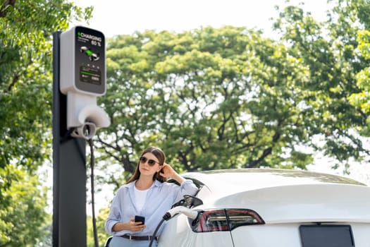 Young woman using smartphone online banking application to pay for electric car battery charging from EV charging station during vacation holiday road trip at national park or summer forest. Exalt