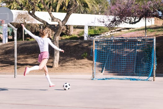 rear view of an unrecognizable young caucasian woman kicking a soccer ball into the goal on an urban field, concept of sport and football lifestyle