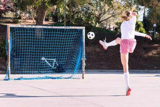 rear view of an unrecognizable young caucasian woman kicking a soccer ball into the goal on an urban field, concept of sport and football lifestyle