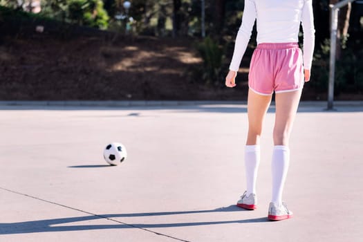 rear view of the legs of an unrecognizable young female football player preparing to kick a soccer ball on an urban field, concept of sport and active lifestyle