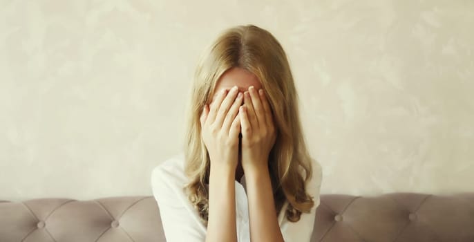 Upset sad crying woman covering her eyes and face with her hands while experiencing mental or physical suffering, fatigue sitting on the couch in room at home