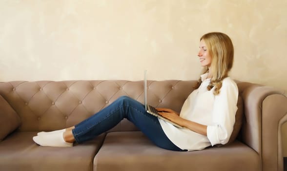 Happy modern young woman working on a laptop sitting on a couch in the living room at home