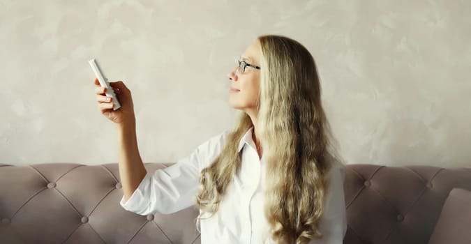 Mature caucasian woman turns on the air conditioner or TV using the remote control sitting on couch in room at home