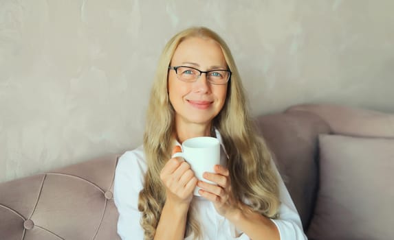 Happy satisfied mature woman enjoying hot tasty cup of coffee or tea looking at camera in early morning sitting on a couch in the living room at home