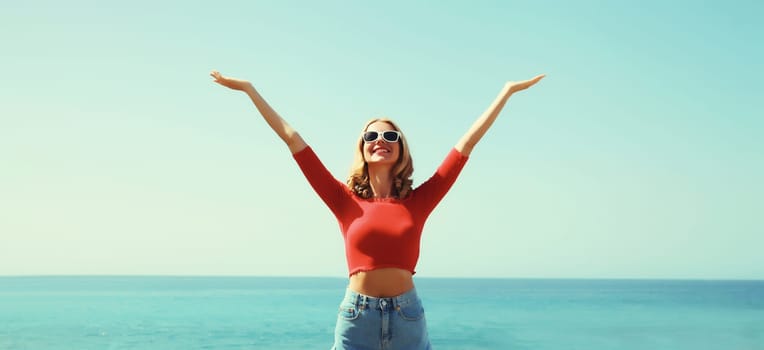 Summer vacation, happy inspired young woman raising her hands up on the beach on sunny sea coast and blue sky background