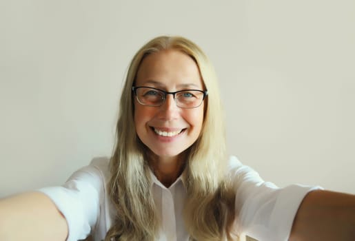 Close up portrait of happy cheerful caucasian mature woman in eyeglasses stretching her hands for taking selfie with mobile phone in office at work or home