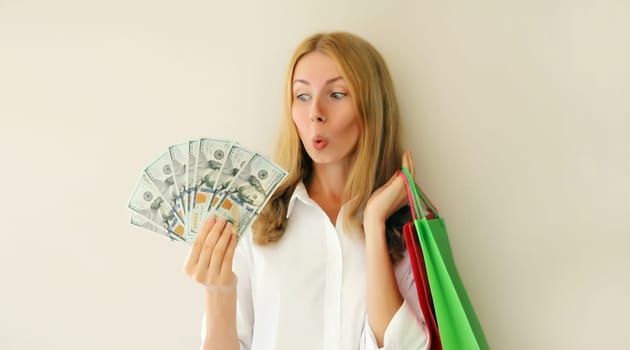 Portrait of surprised shocked young woman with shopping bags holding cash money looking at dollar bills on white background