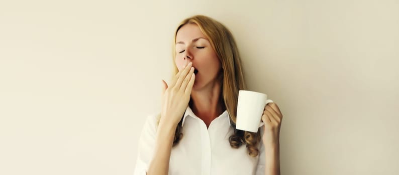Sleepy lazy young woman yawns waking up after sleeping holds cup of coffee in morning on white background