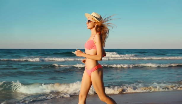Summer vacation, happy slim woman walking running in bikini swimsuit and tourist straw hat on the beach on sea coast with waves background on sunny day