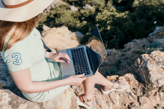 Digital nomad, woman in the hat, a business woman with a laptop sits on the rocks by the sea during sunset, makes a business transaction online from a distance. Freelance, remote work on vacation.