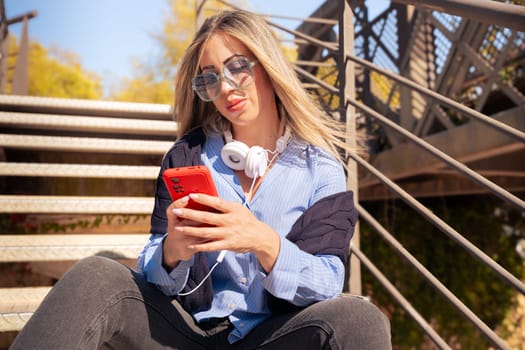 Portrait of a smiling young woman wearing headphones and sunglasses, using a social media app