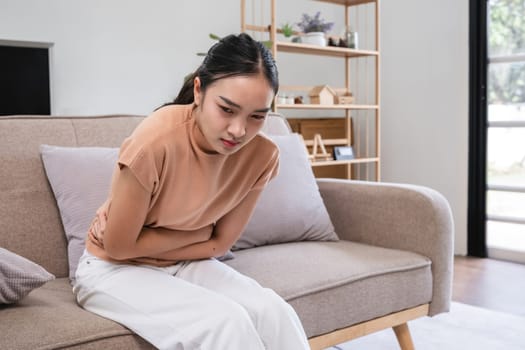 Young woman with stomach pain from menstruation sitting on a sofa, holding her abdomen, in a modern living room. Health and wellness concept.