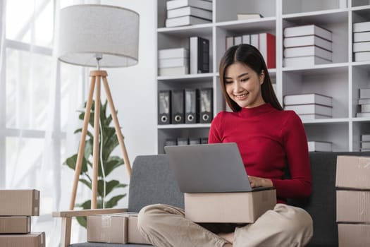 Young female business owner managing online sales from home, using a laptop and surrounded by packages, representing small business and e-commerce entrepreneurship.