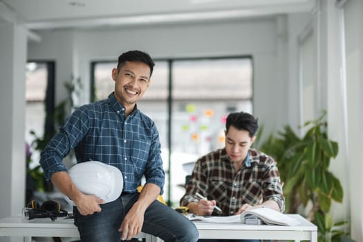 Two male engineers working together in a modern office, reviewing blueprints and wearing safety gear, highlighting teamwork and professionalism in the engineering field.