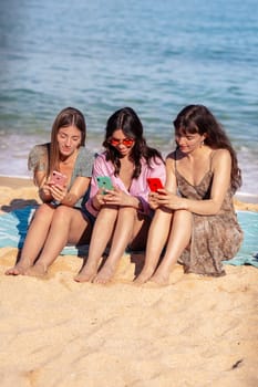 Young Happy Caucasian Friends On Vacation Using Smartphone App Sitting On Towels On Beach