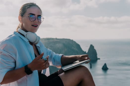 Digital nomad, woman in the hat, a business woman with a laptop sits on the rocks by the sea during sunset, makes a business transaction online from a distance. Freelance, remote work on vacation.