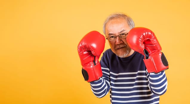 Portrait Asian old man wearing glasses wear two red boxing gloves studio shot isolated yellow background, smiling happy elderly man gray haired healthy fighter lifestyle concept