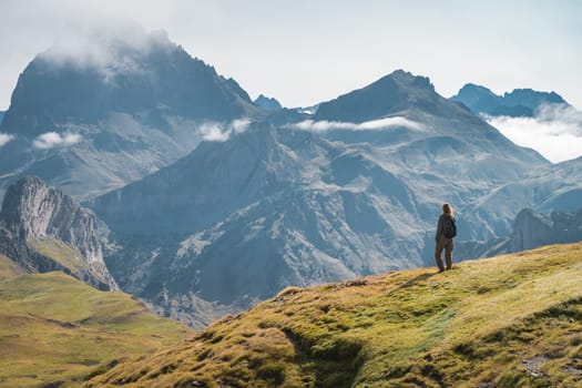 A lone hiker marvels at rugged mountains under a clear sky with scattered clouds on an alpine expedition