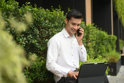 Smiling asian male entrepreneur sitting outdoor talking on mobile phone and using digital tablet.