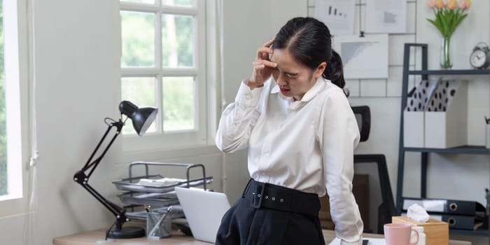 Businesswoman in office suffering from headache, surrounded by documents, laptop, and office supplies, depicting workplace stress.