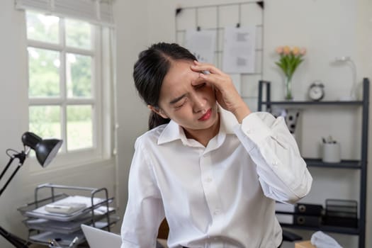 A businesswoman in a white shirt experiencing stress and headache in an office setting, surrounded by documents and a laptop.