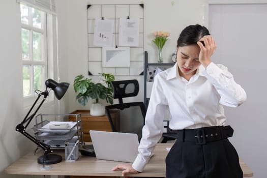 A businesswoman in a white shirt and black pants experiencing a headache in a modern office setting with a laptop and documents.