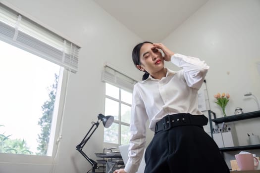 A businesswoman in a modern office environment experiencing stress and headache, standing near a desk with natural light coming through the window.