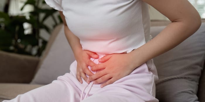 A woman in casual clothing sitting on a couch, holding her stomach in pain, indicating discomfort or digestive issues.